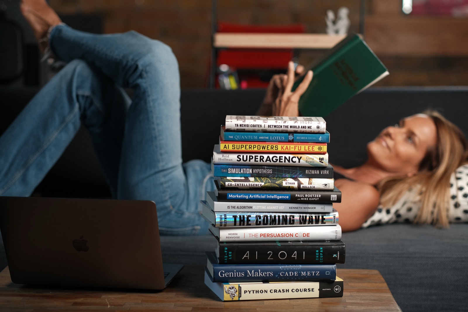 Angie Carel reading, beside a tall stack of AI books and a laptop